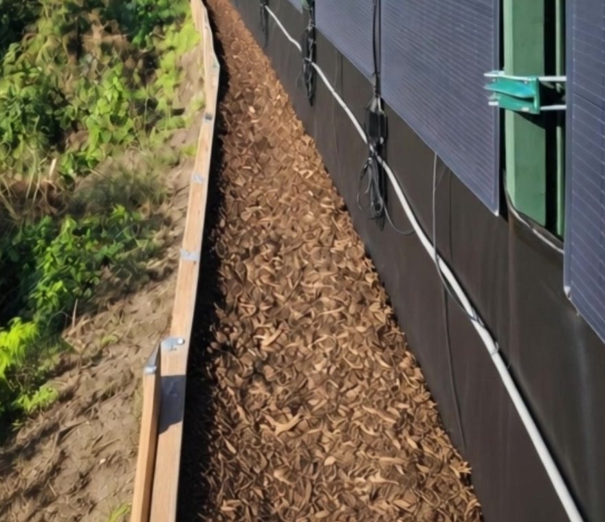 Flexible solar panels held by brackets on green steel fence posts above a woodchip path